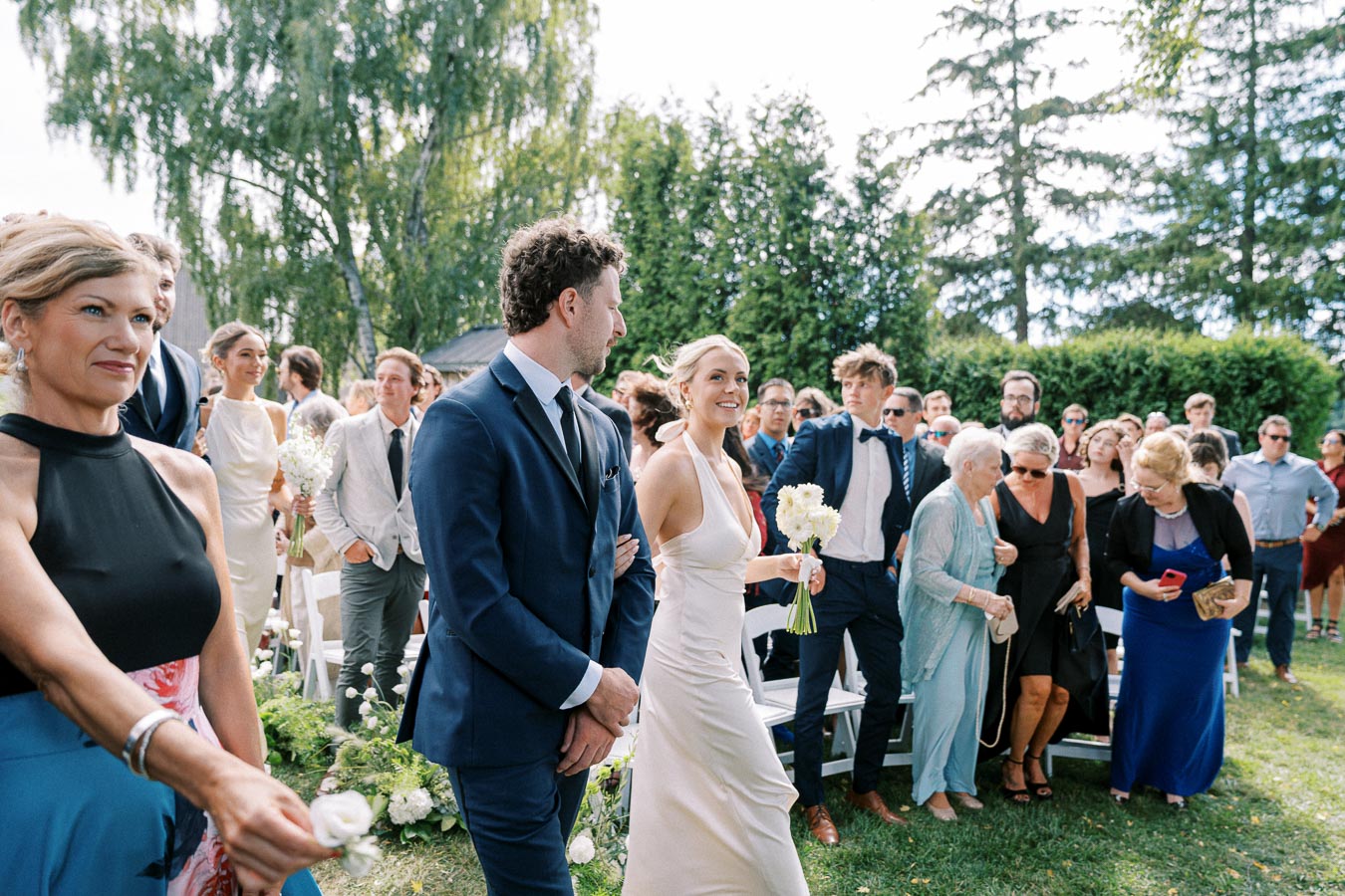 Wedding procession with bride and groom walking through a garden setting surrounded by guests. The bride carries white flowers, and people are dressed in formal attire, celebrating on a sunny day with greenery in the background.
