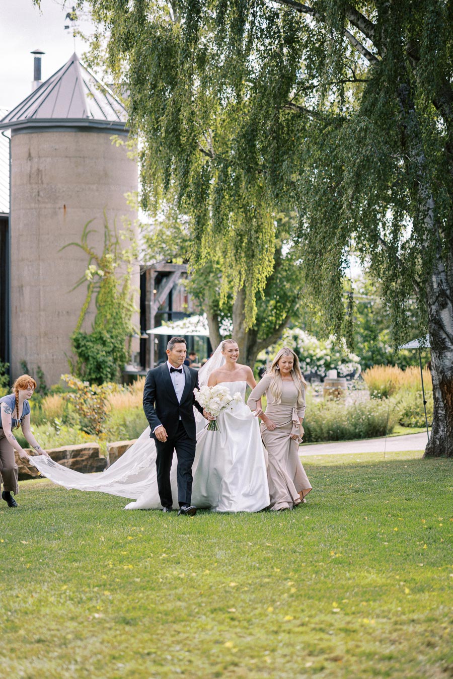 A bride in a white wedding dress walks down a grassy aisle accompanied by two attendants, with a picturesque outdoor venue in the background, including lush greenery and a rustic building.