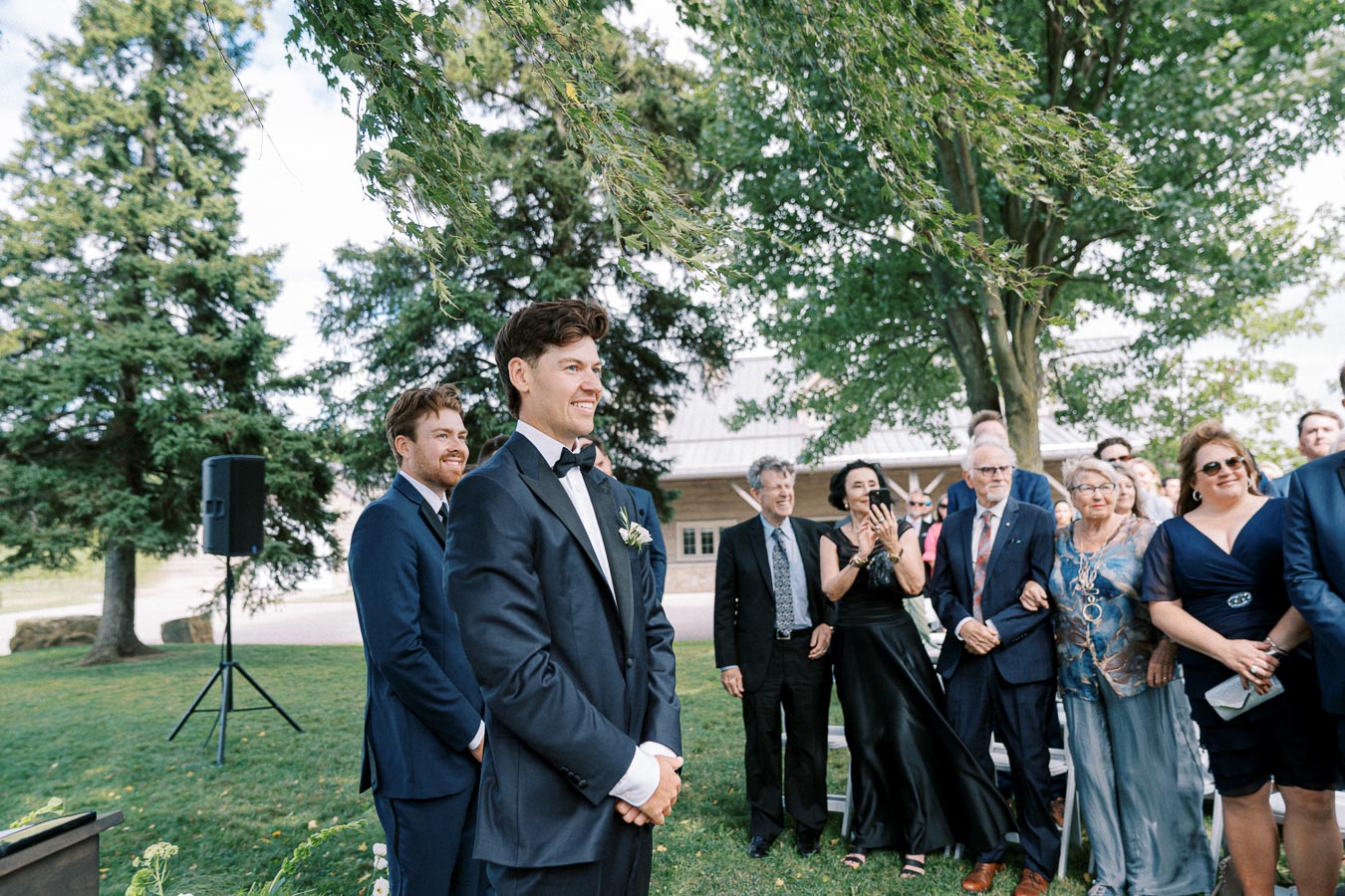 A groom in a navy suit with a boutonniere smiles while standing outdoors during a wedding ceremony, surrounded by guests in formal attire under trees and clear skies.