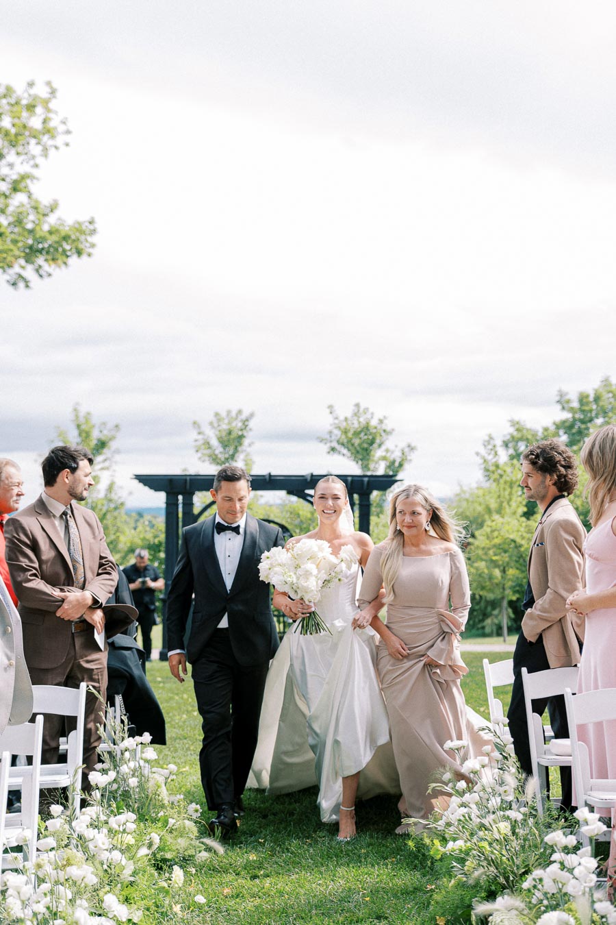 A bride in a white gown walks down an outdoor aisle with her parents, surrounded by guests, green trees, and white flowers, on a sunny wedding day.