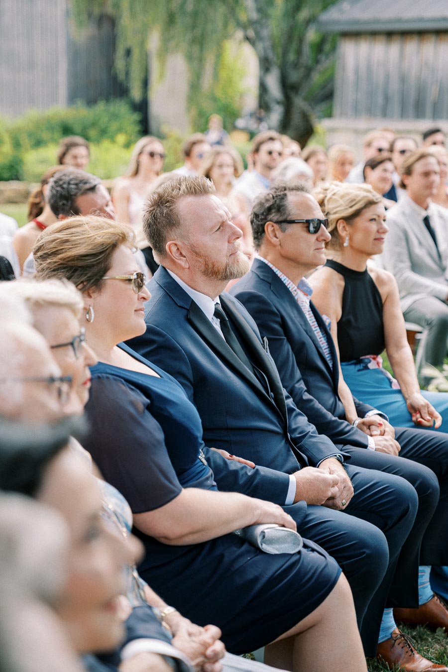 Audience attentively seated outdoors in formal attire during a daytime event, surrounded by lush greenery and rustic wooden buildings.
