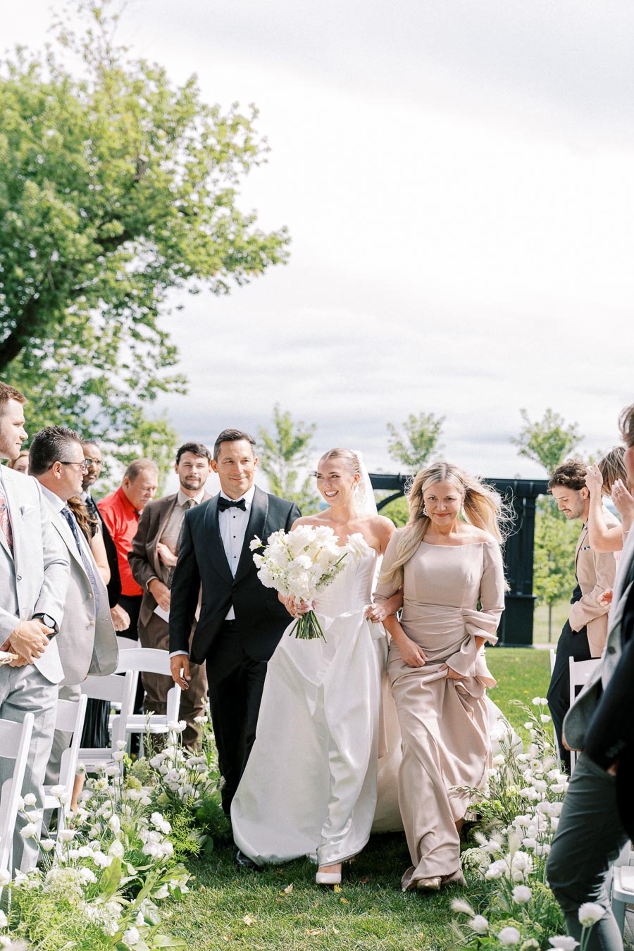 A bride walking down the aisle with her parents during an outdoor wedding ceremony, surrounded by guests and lush greenery. She is holding a bouquet of white flowers and smiling brightly.