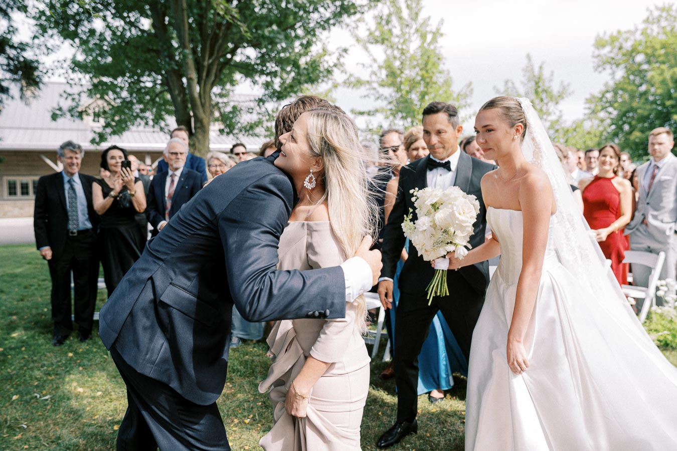A joyous wedding scene with a bride in a white dress holding a bouquet of white flowers, surrounded by elegantly dressed guests. The groom embraces a woman in celebration, while onlookers, including a man in a tuxedo, smile and applaud under a tree-filled setting.