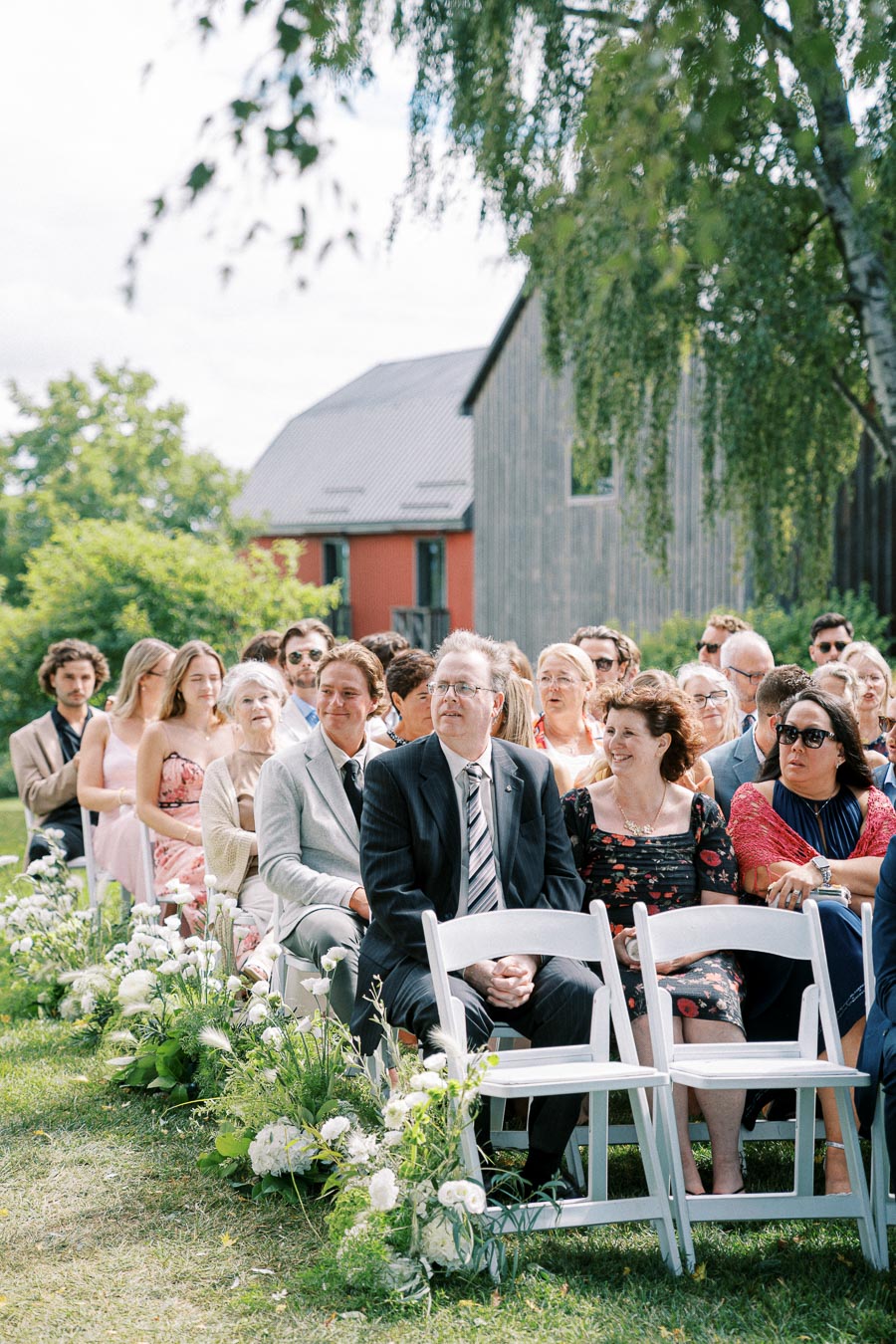 Guests seated outdoors at a wedding ceremony in a garden setting, surrounded by lush greenery and floral arrangements. A rustic barn is visible in the background under a partly cloudy sky.