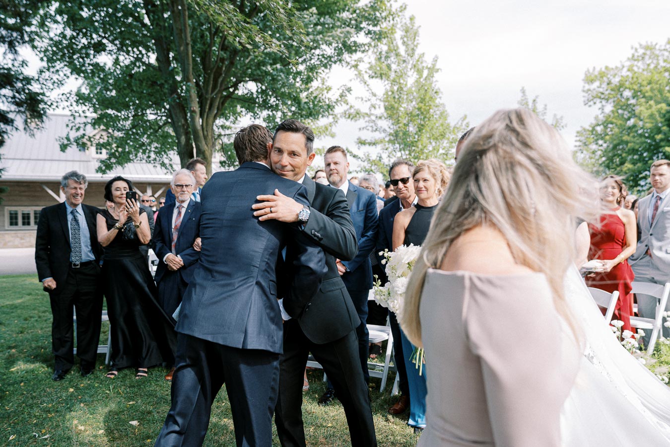 A joyful wedding ceremony outdoors with guests smiling as two men in suits embrace, surrounded by seated people in formal attire under a green, sunny setting.