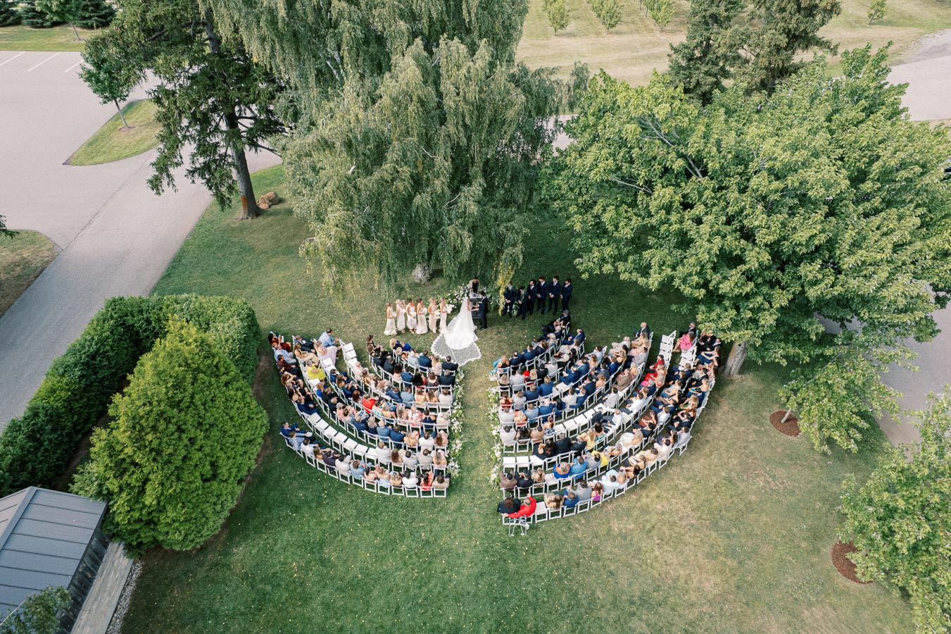 Aerial view of an outdoor wedding ceremony with a bride and groom standing under a large tree, surrounded by guests seated in a semi-circle arrangement, set in a scenic green park.