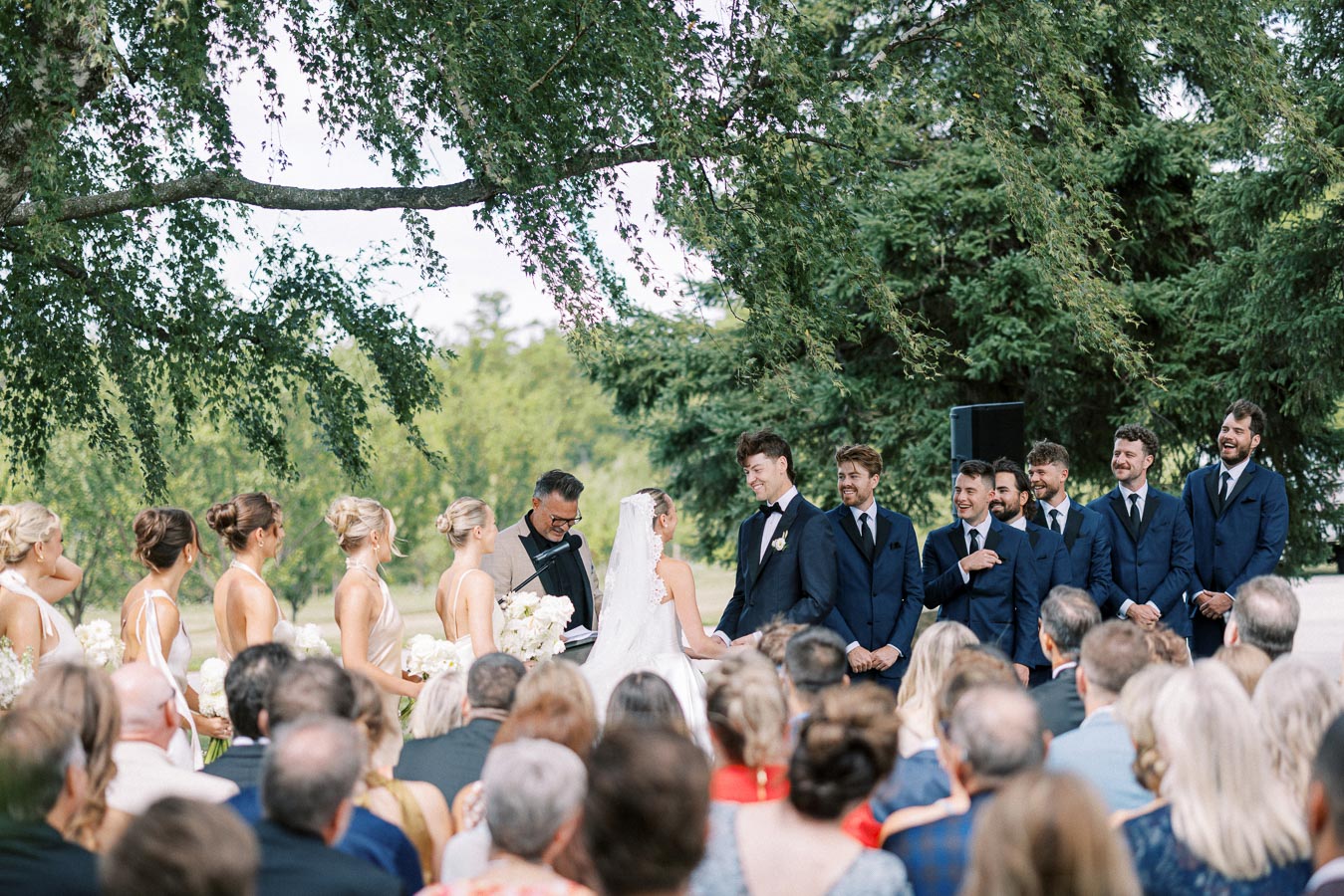 Outdoor wedding ceremony with bride and groom exchanging vows under a tree, surrounded by bridesmaids in white dresses and groomsmen in blue suits. Guests sit attentively, enjoying the beautiful natural setting.