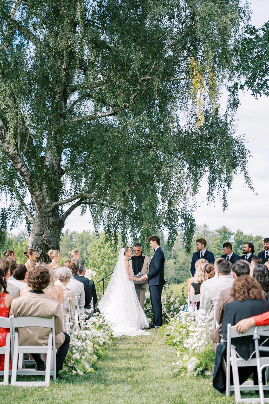 Outdoor wedding ceremony under a large tree, featuring a bride and groom exchanging vows surrounded by guests seated in rows, with floral arrangements along the aisle and a lush, green backdrop.