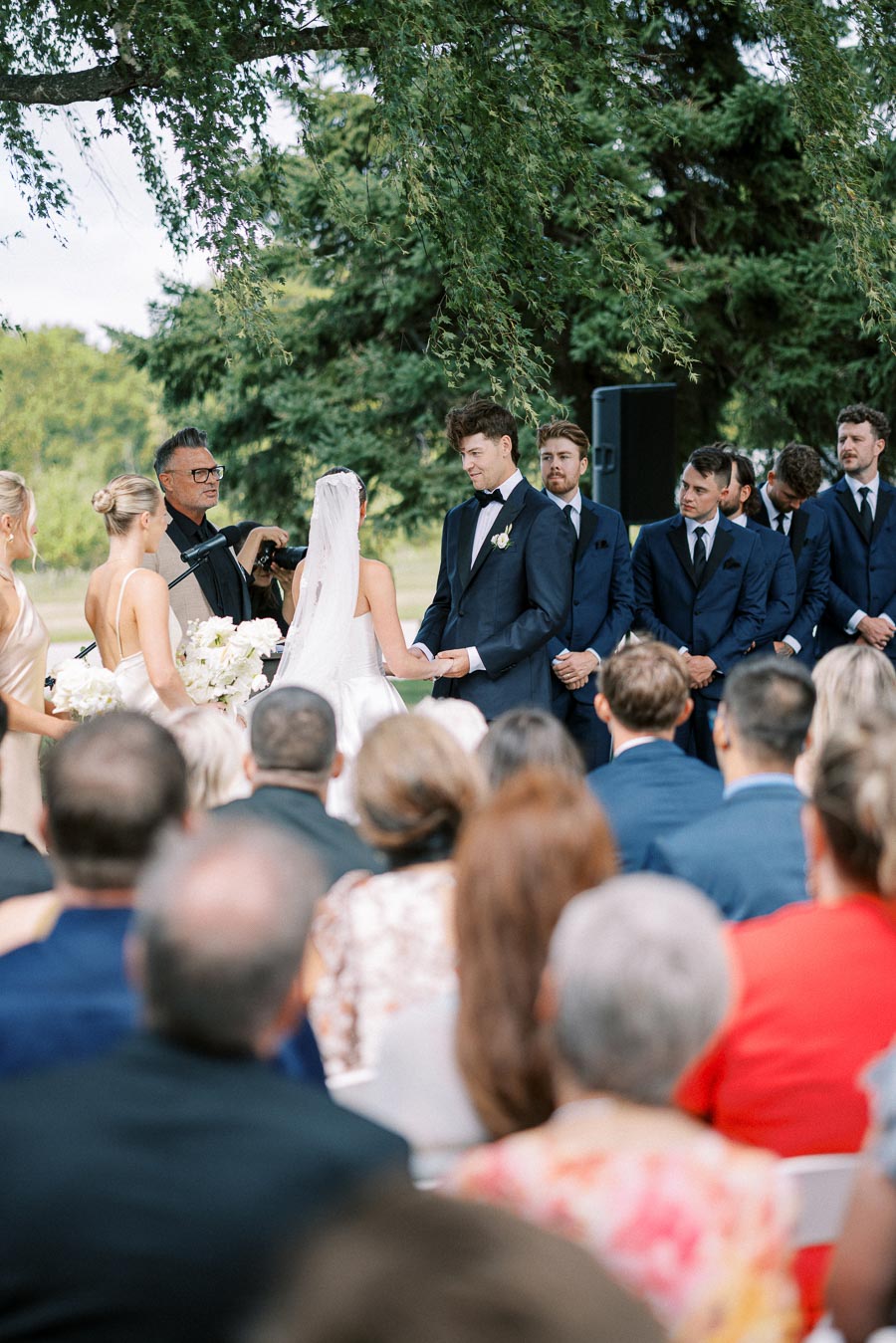 Outdoor wedding ceremony with a bride in a white gown and veil, groom in a dark suit, bridesmaids in light dresses, and groomsmen in dark suits, set under a tree with guests seated.