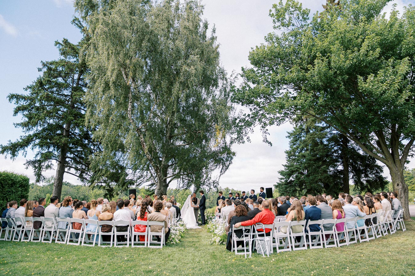 Outdoor wedding ceremony under trees with seated guests and a couple exchanging vows.