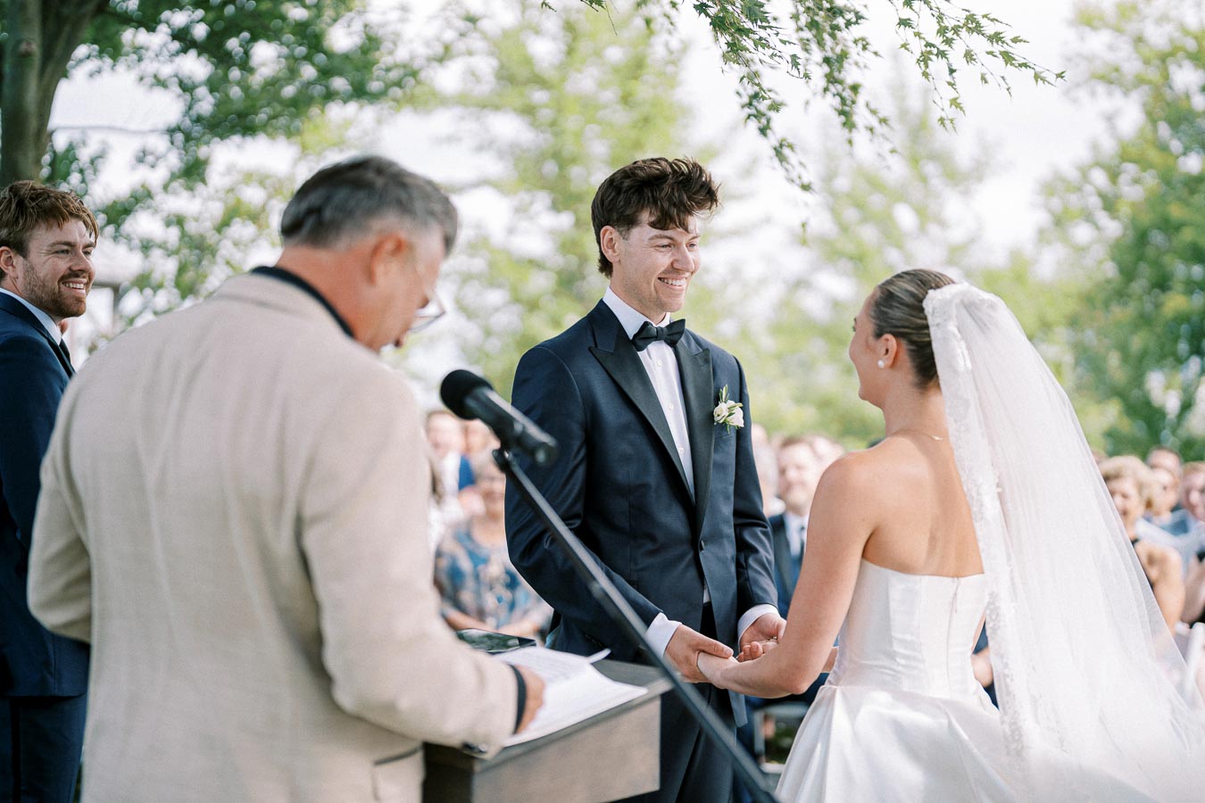 A bride and groom exchange vows during an outdoor wedding ceremony, with the officiant speaking into a microphone and a guest smiling in the background. The groom is in a dark suit, and the bride is wearing a white gown with a veil, surrounded by trees.