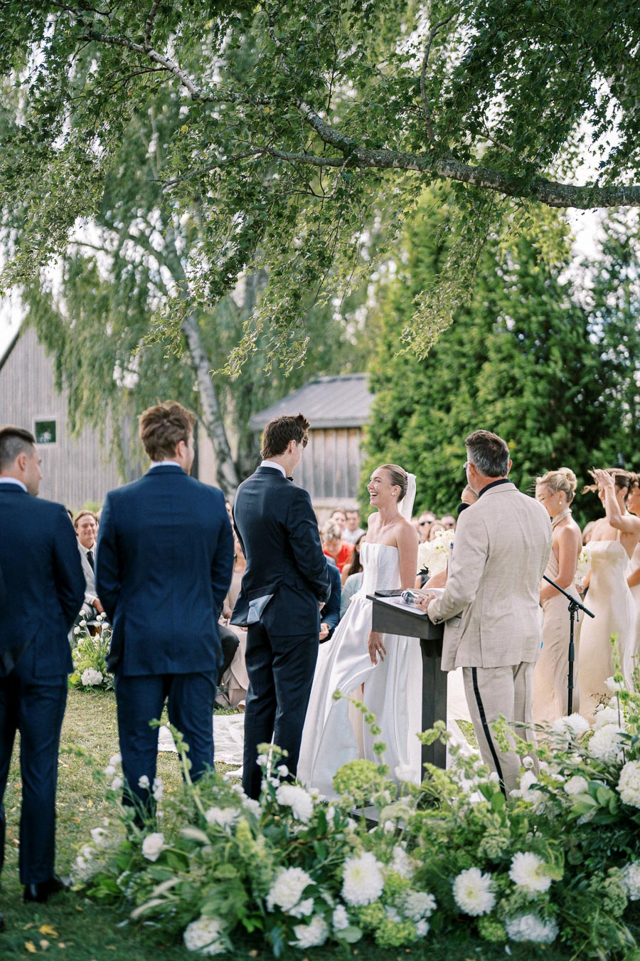 Outdoor wedding ceremony with a bride and groom exchanging vows under a tree, surrounded by guests and bridesmaids, featuring lush greenery and floral decorations.