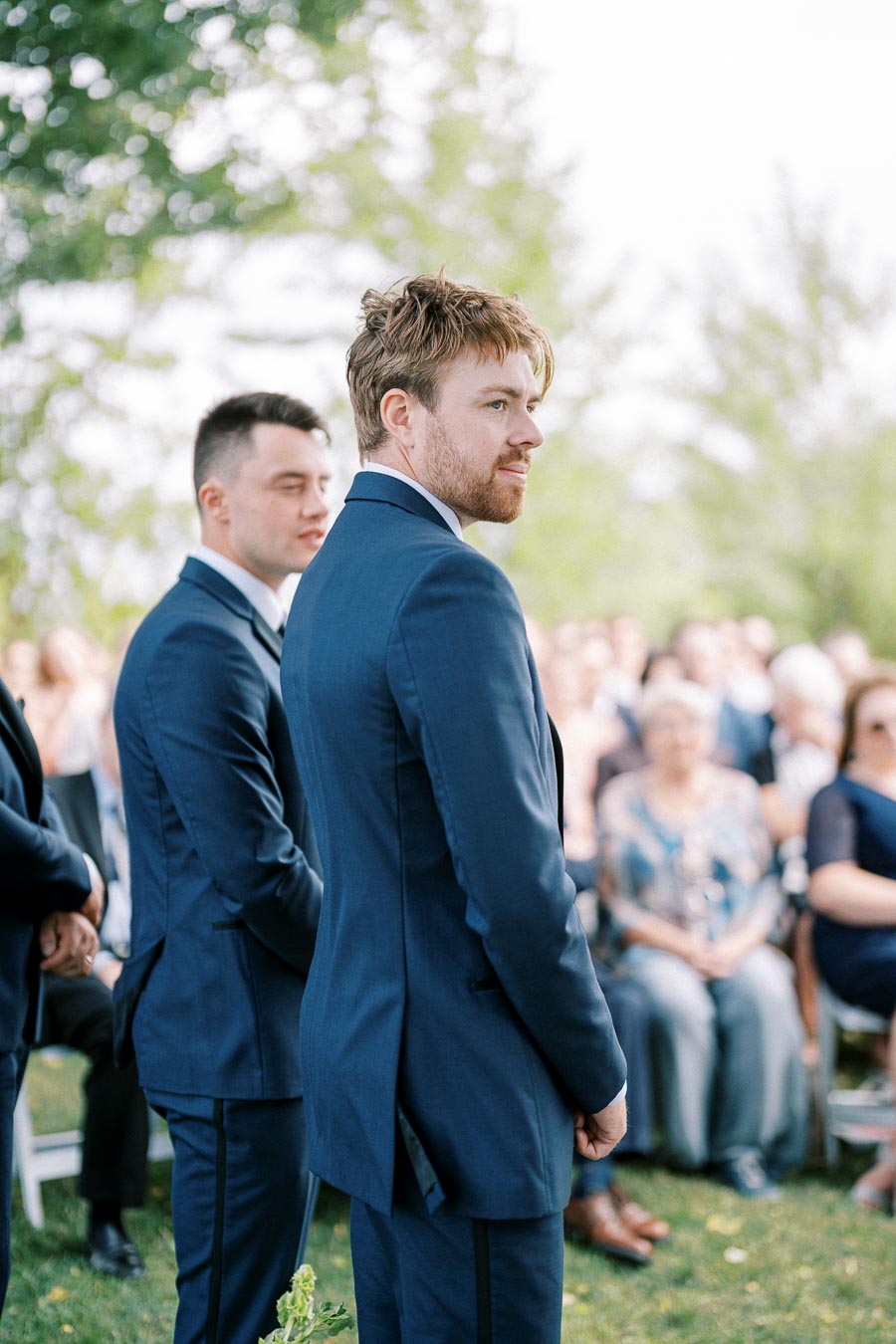 Groomsmen in blue suits stand outdoors at a wedding ceremony, with guests seated in the background, surrounded by greenery.