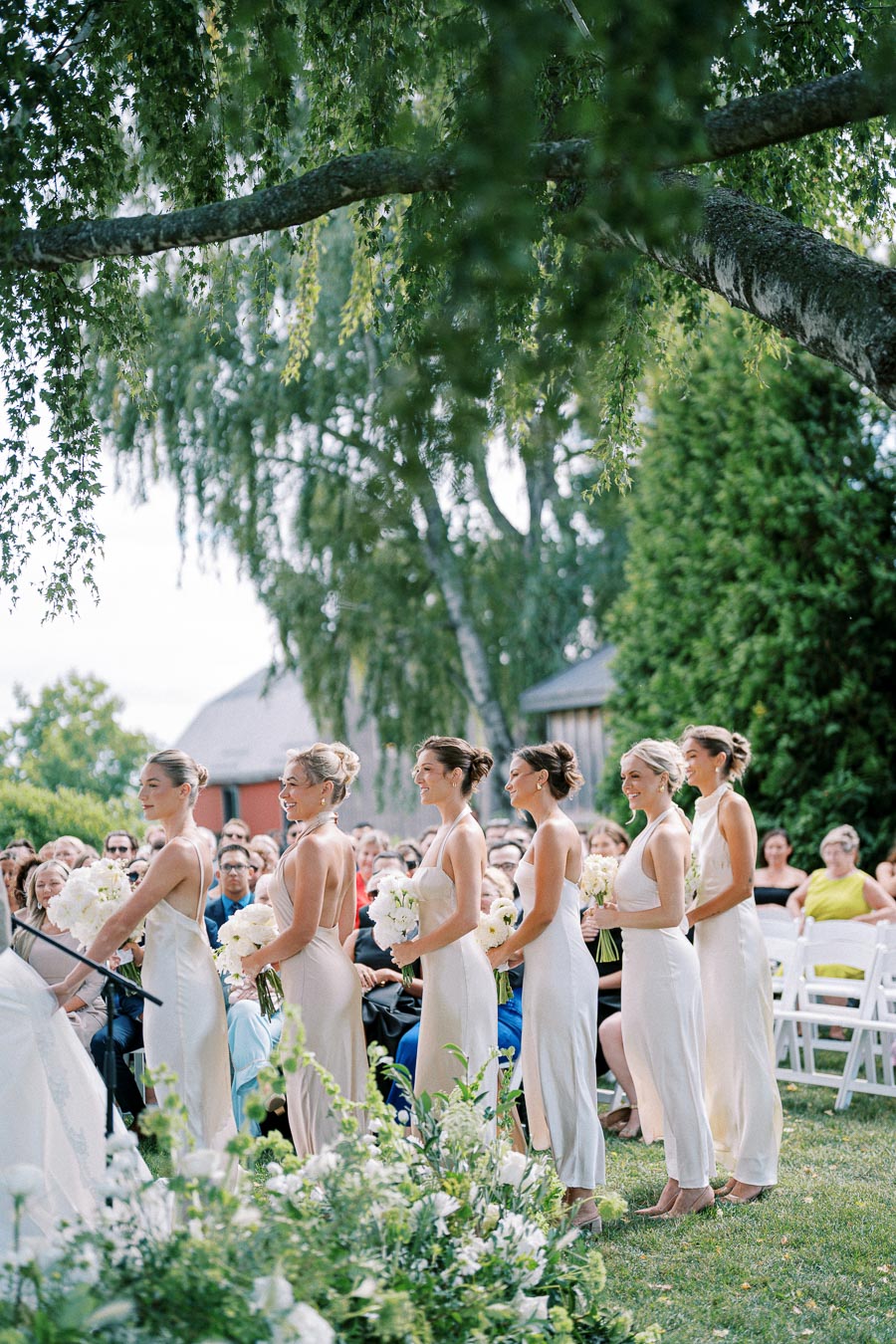 Six bridesmaids in matching dresses stand in a garden setting, holding white flower bouquets, surrounded by guests seated on white chairs. Lush greenery and an elegant floral arrangement enhance the outdoor wedding ceremony.