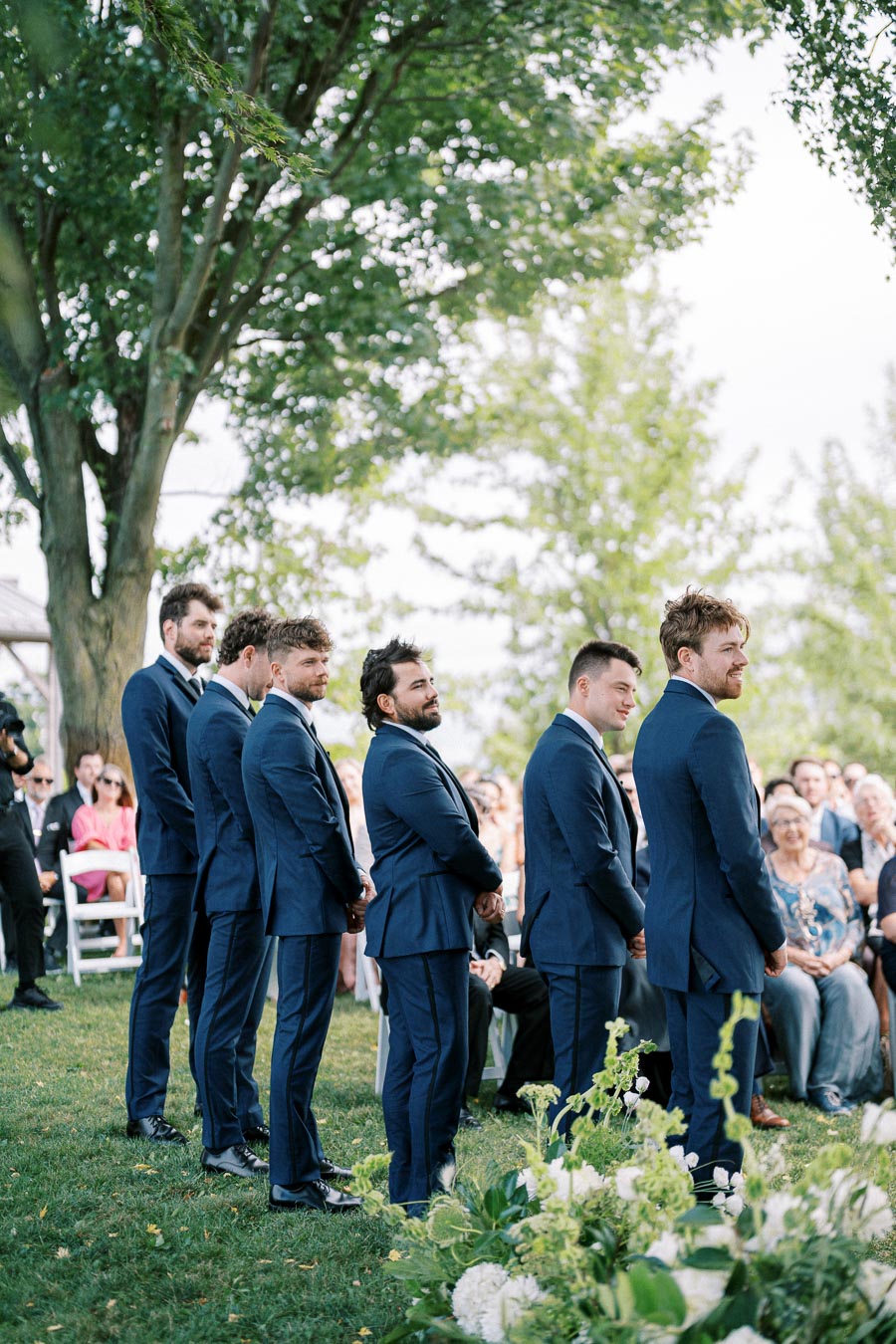 Groomsmen in blue suits standing in a line during an outdoor wedding ceremony, surrounded by greenery and seated guests.