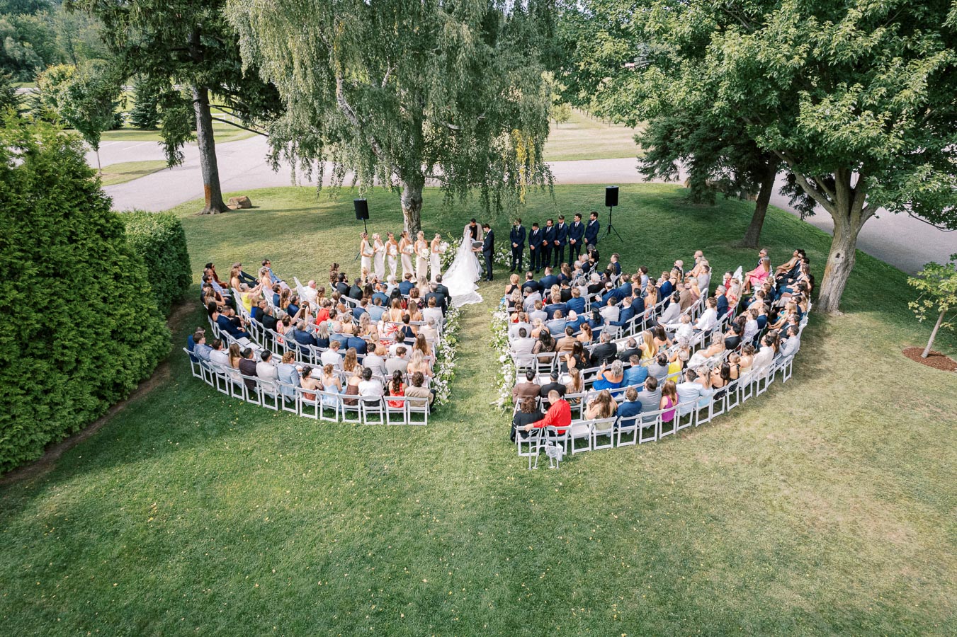 Aerial view of an outdoor wedding ceremony in a lush garden setting, featuring a bride and groom under a large tree, surrounded by guests seated in a semicircular arrangement with bridesmaids and groomsmen standing nearby.