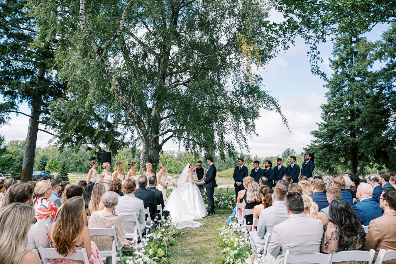 Outdoor wedding ceremony with a couple exchanging vows under a large tree, surrounded by bridesmaids in light dresses and groomsmen in dark suits, with guests seated in white chairs.