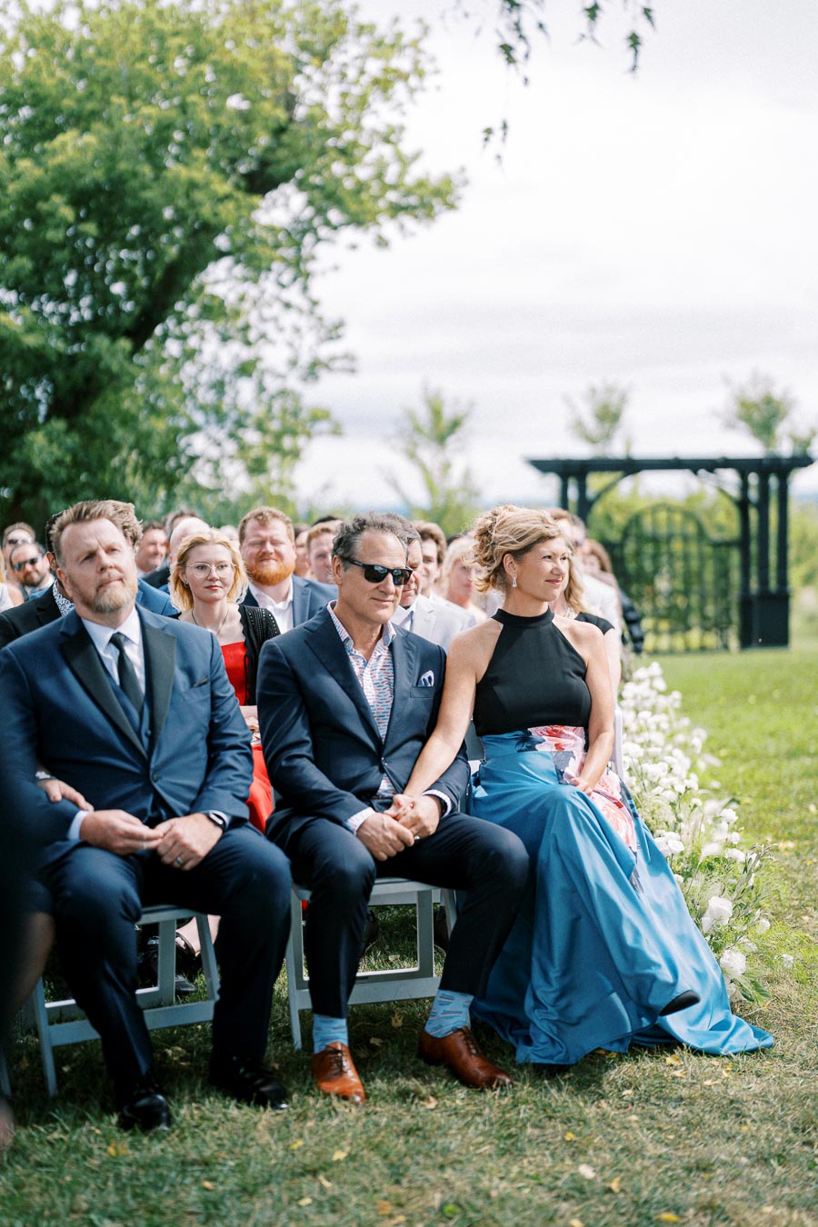 Elegant outdoor wedding ceremony with guests dressed in formal attire, seated in rows on a lush green lawn with floral arrangements and trees in the background.