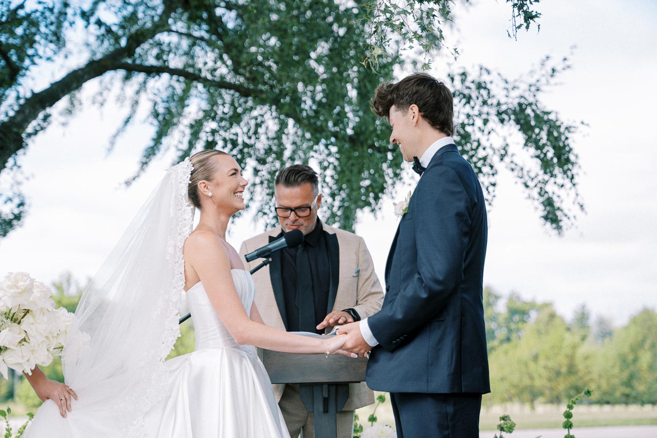 A bride in a white wedding dress and a groom in a navy suit hold hands during an outdoor wedding ceremony, with an officiant in the background under a large tree.