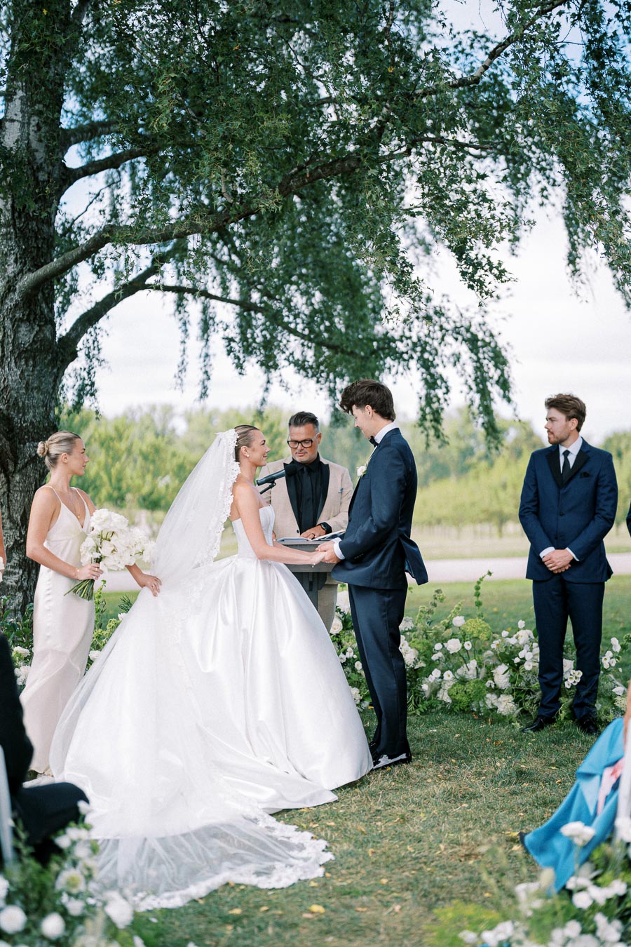 Wedding ceremony outdoors under a large tree, featuring a bride in a white gown, groom in a dark suit, officiant, and bridesmaid holding flowers.