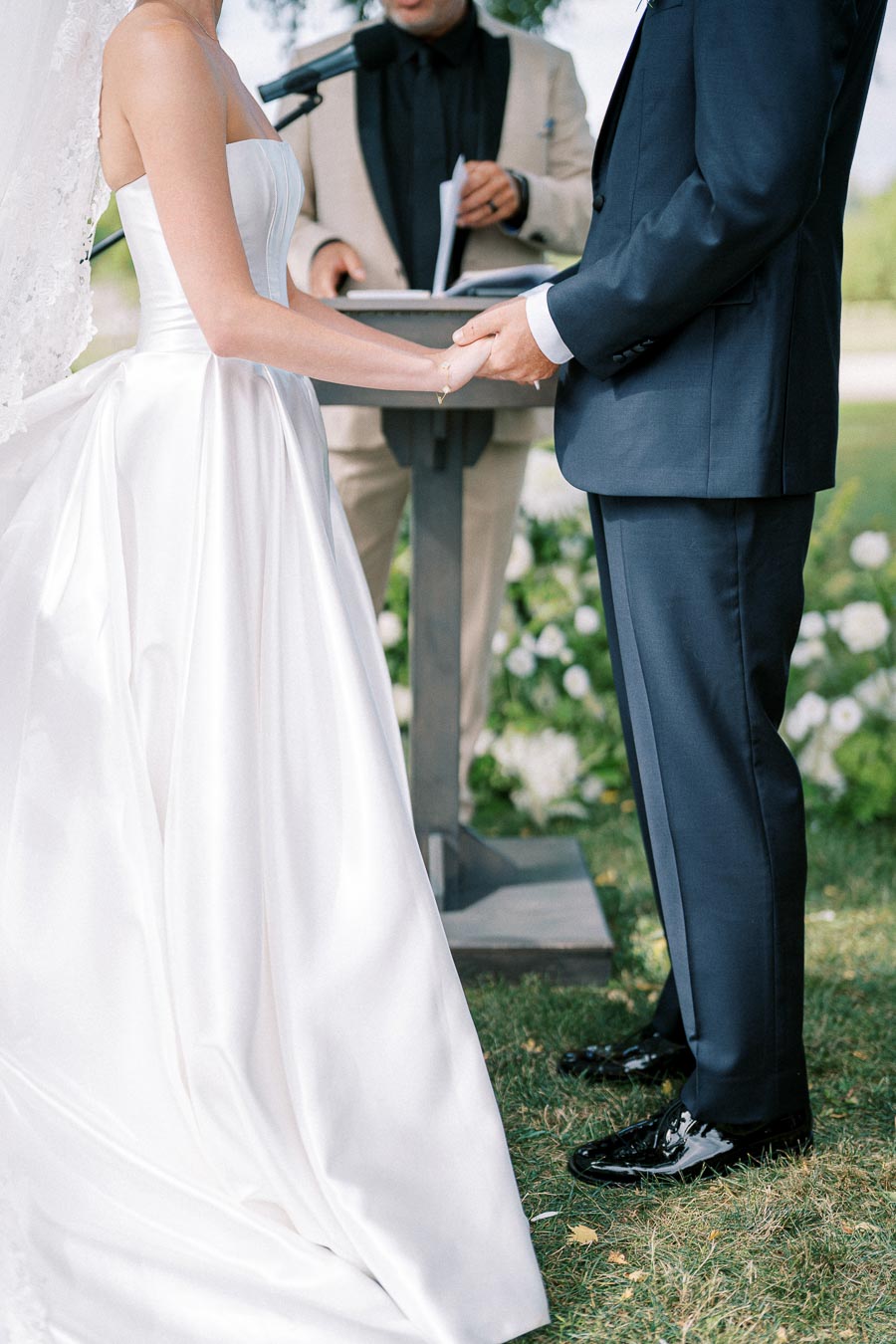 Elegant outdoor wedding ceremony with bride in white gown and groom in dark suit exchanging vows, officiant in the background.
