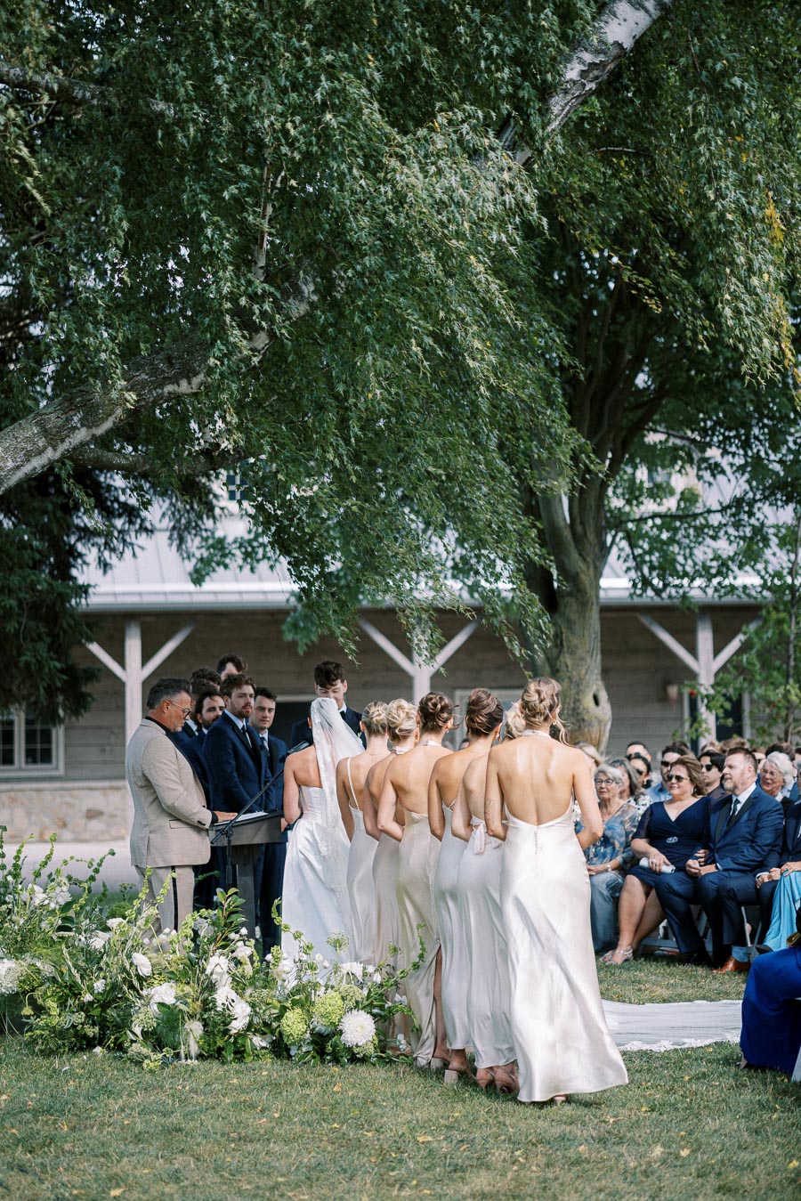 Outdoor wedding ceremony with bridesmaids in matching elegant dresses standing beside the bride under a tree, surrounded by guests and floral decorations.