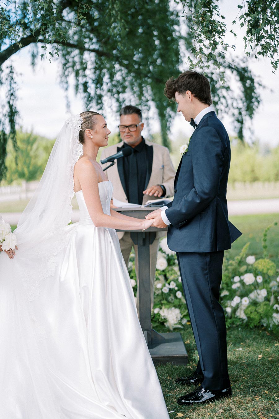 Bride and groom exchanging vows outdoors under a lush tree, with an officiant conducting the wedding ceremony; the bride wears a flowing white gown and veil, and the groom is in a blue suit.