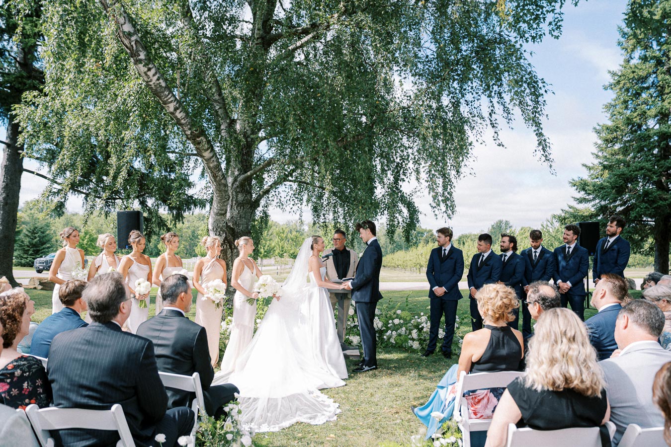 Outdoor wedding ceremony with a bride in a white gown and groom in a dark suit exchanging vows under a large tree, surrounded by bridesmaids in light dresses and groomsmen in blue suits, sunny garden setting.