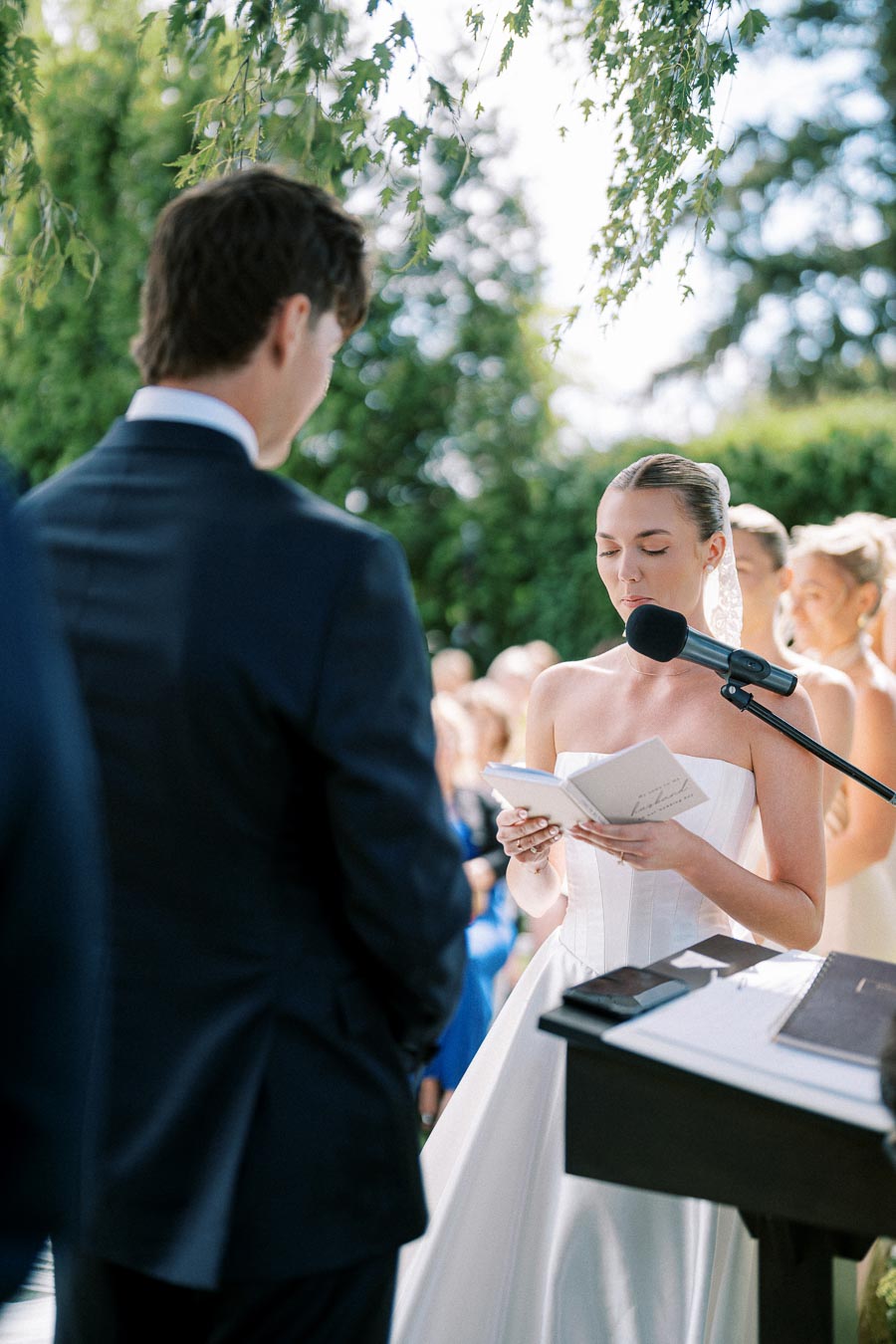 Bride reading vows at outdoor wedding ceremony, wearing a strapless white gown and lace veil, with groom in a dark suit. Microphone and trees in the background enhance the romantic, natural setting.
