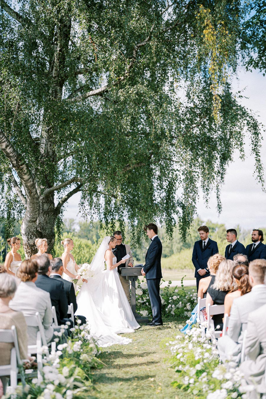 Outdoor wedding ceremony with a bride and groom exchanging vows under a large tree, surrounded by bridesmaids, groomsmen, and seated guests on a sunny day.