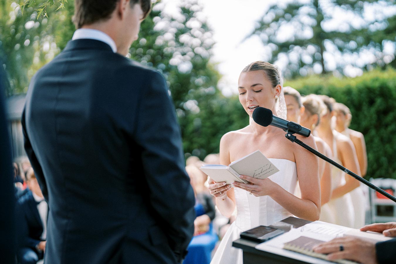 A bride reads her vows at an outdoor wedding ceremony, holding a microphone and a booklet, with bridesmaids in the background, under a clear sky surrounded by greenery.