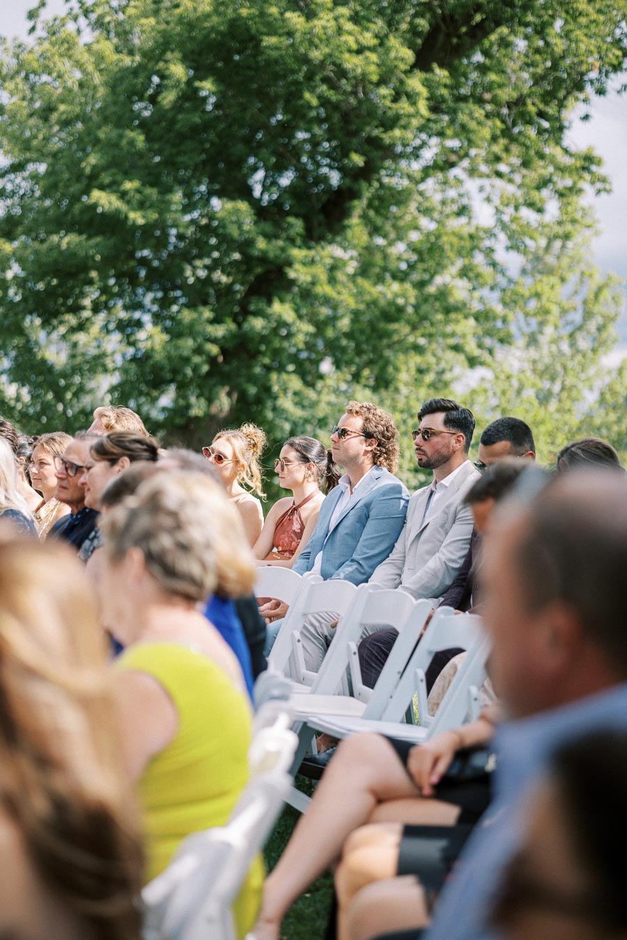 Group of people seated outdoors at a sunny event, dressed in summer attire and sunglasses, with lush green trees in the background.