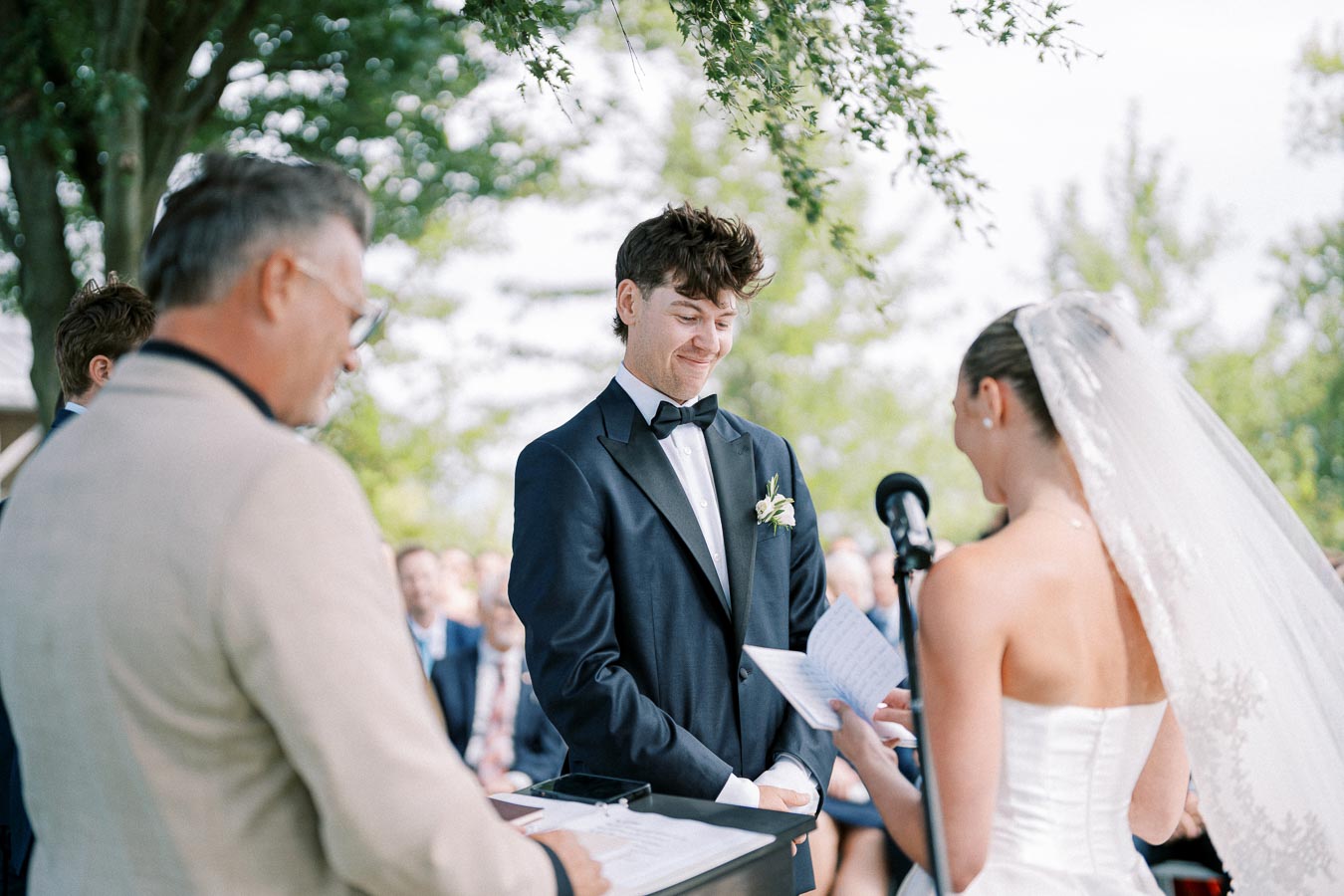 Outdoor wedding ceremony with a bride and groom exchanging vows, officiant standing nearby, surrounded by guests on a sunny day.