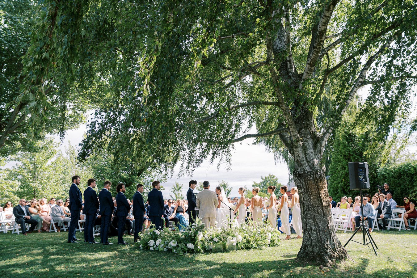 Outdoor wedding ceremony under a large tree, featuring a bride, groom, bridesmaids, and groomsmen in elegant attire, surrounded by guests seated on white chairs amidst lush greenery and floral arrangements.