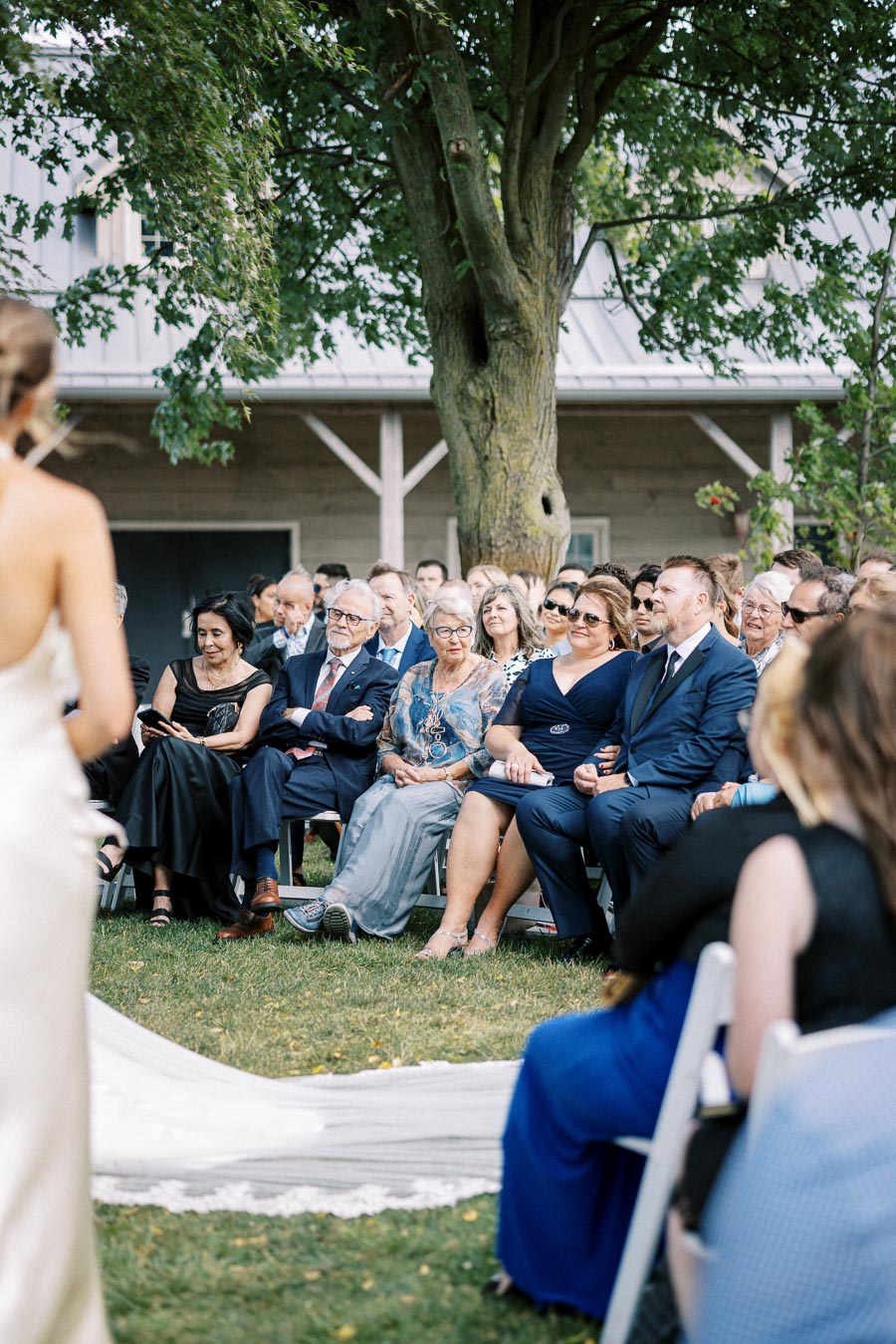 Guests seated at an outdoor wedding ceremony under a large tree, dressed in formal attire, attentively watching the event unfold.