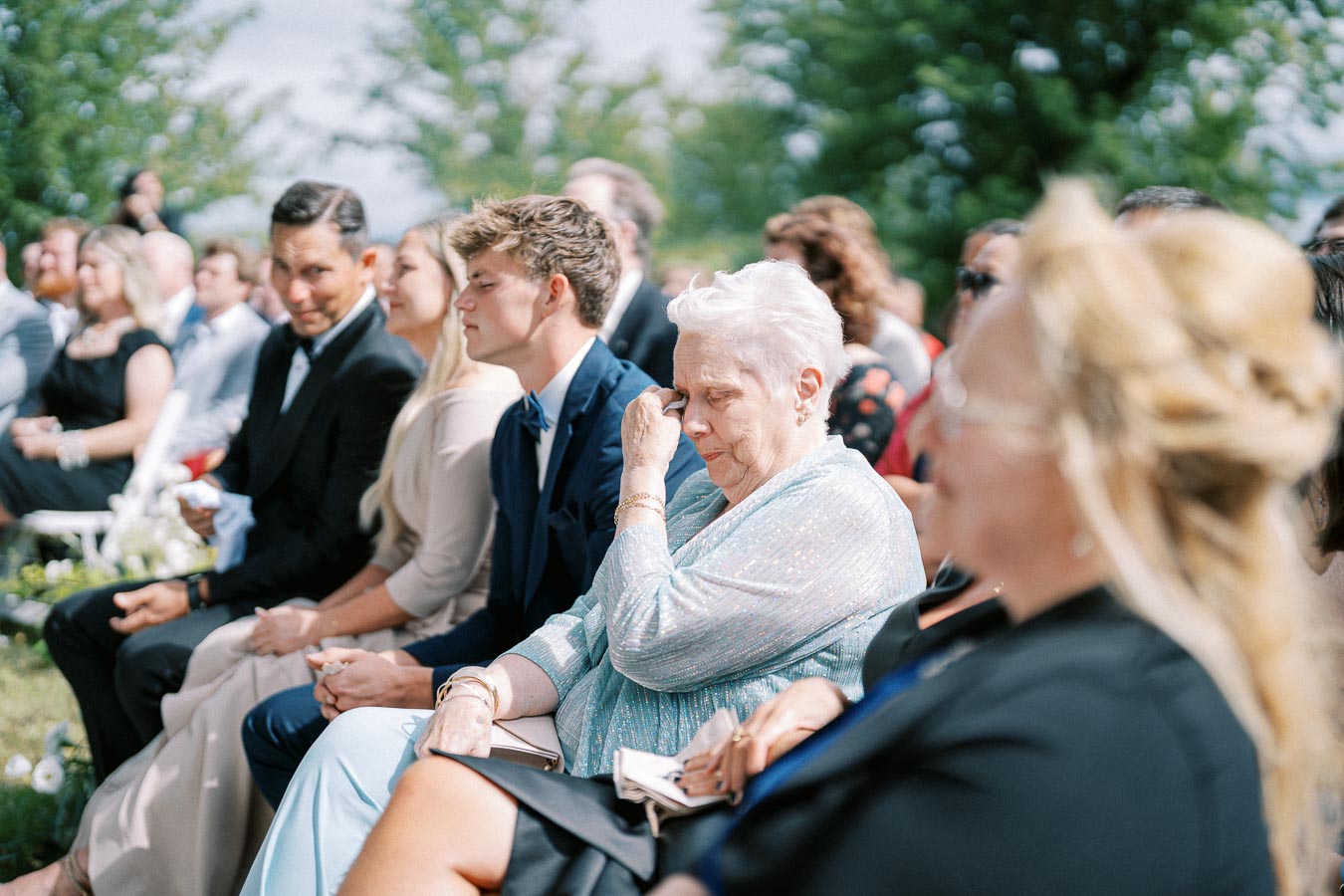 A group of people seated outdoors at a daytime event, with a woman in a light blue outfit wiping her eyes, capturing an emotional moment. The setting features trees and greenery in the background, suggesting a natural, serene atmosphere.