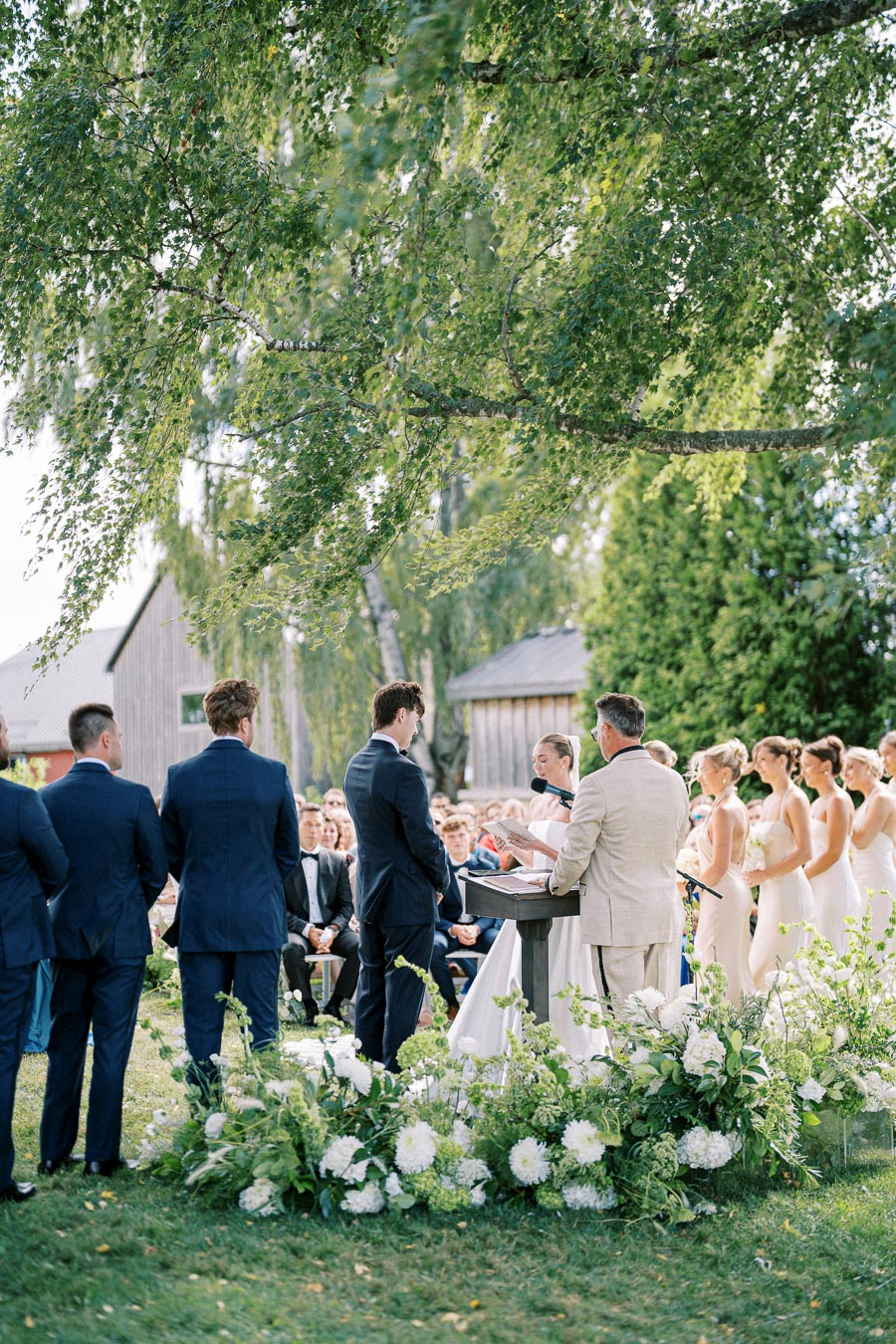 Outdoor wedding ceremony under a large tree with a couple exchanging vows, surrounded by bridesmaids in white dresses and groomsmen in blue suits.