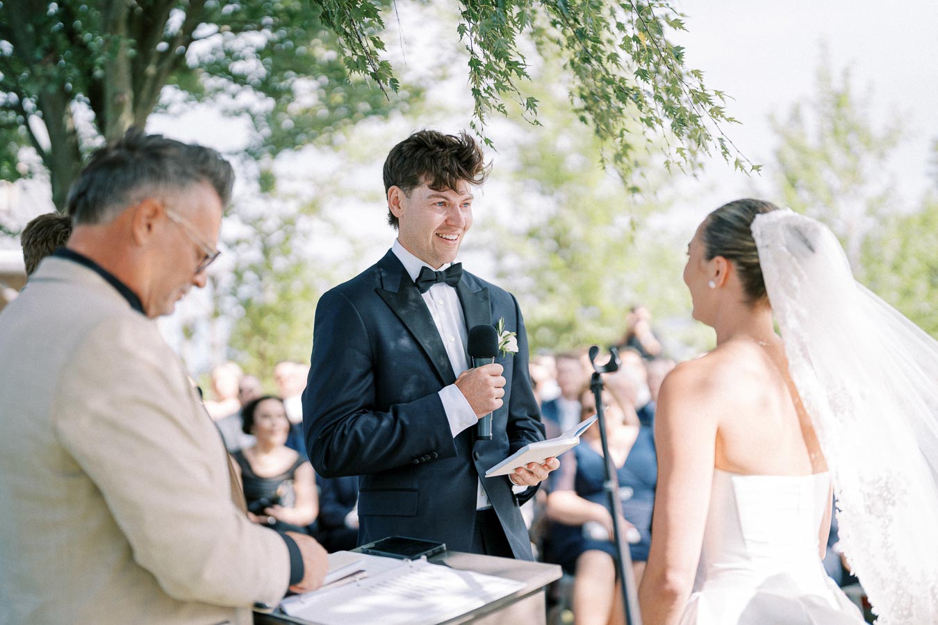 A groom in a navy tuxedo reads his vows to the bride, who is wearing a white wedding dress with a veil, during an outdoor wedding ceremony under a tree. An officiant stands nearby with a notepad, while guests watch attentively in the background.