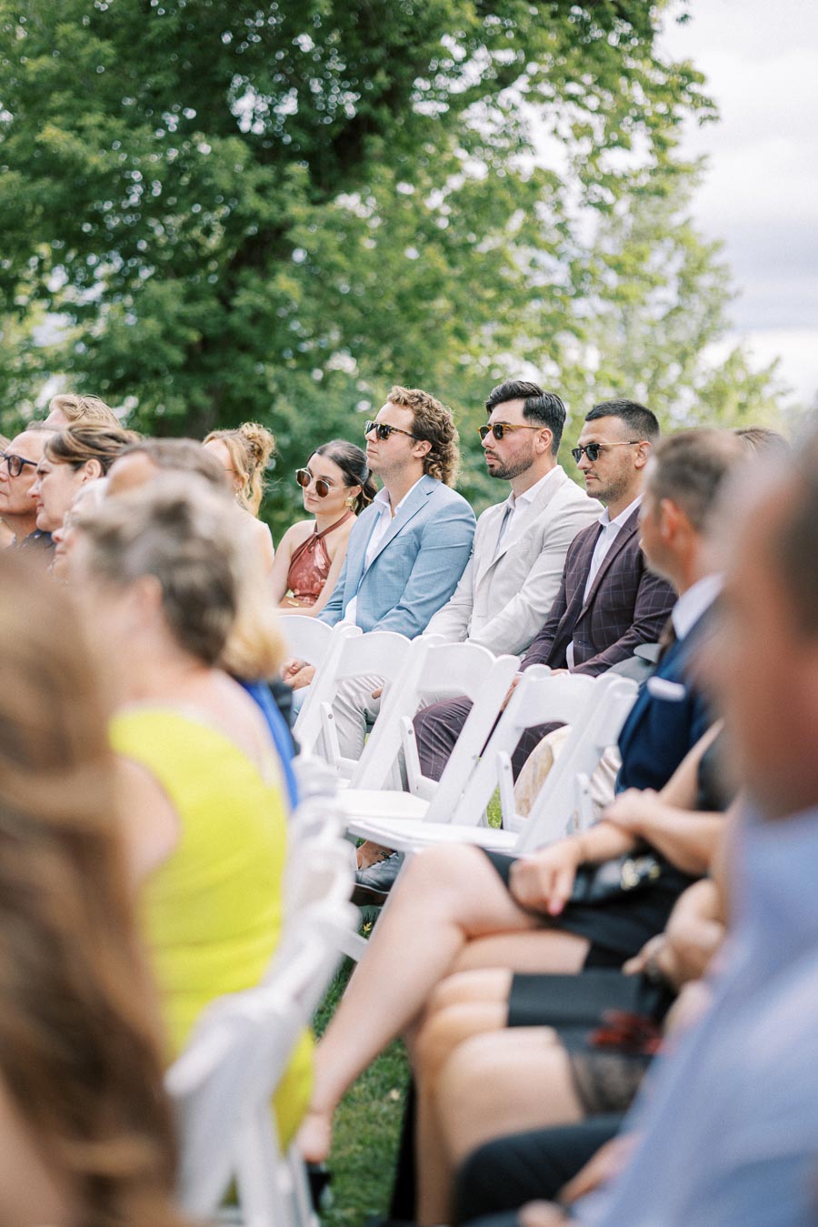 A group of stylishly dressed people sitting outdoors, attentively watching a ceremony, with lush green trees in the background.