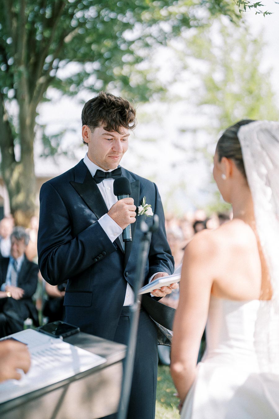 A groom in a navy tuxedo reads vows to the bride during an outdoor wedding ceremony, with a microphone in hand and guests seated in the background on a sunny day.