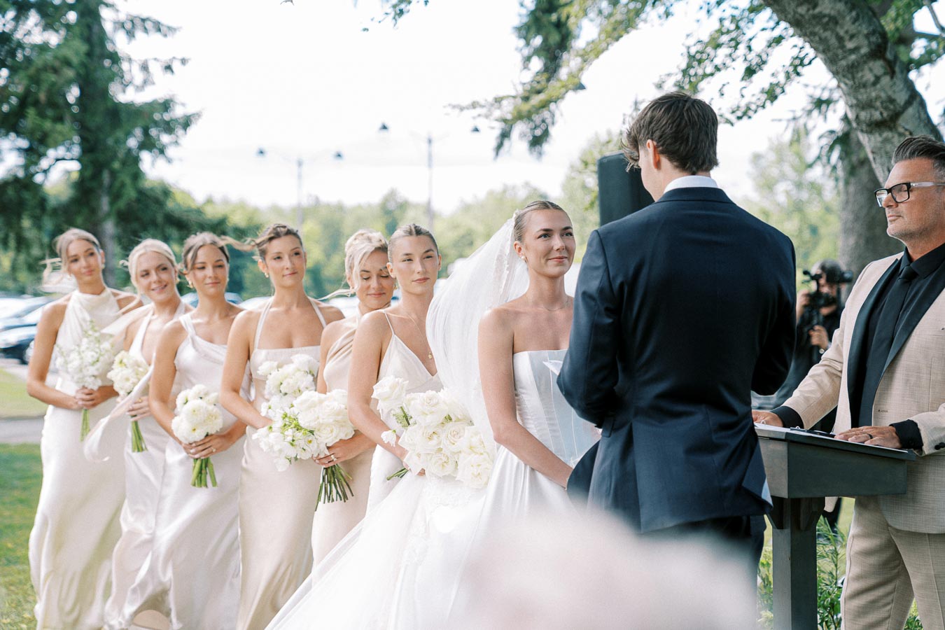 Wedding ceremony scene with a bride and groom exchanging vows outdoors, surrounded by bridesmaids in light dresses holding white floral bouquets, all set against a backdrop of lush greenery and a ceremonial officiant.