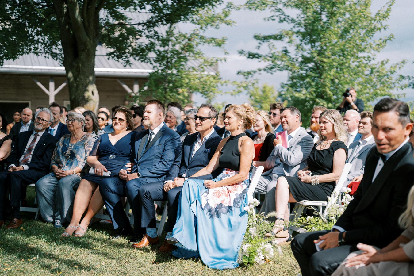 A group of well-dressed guests seated outdoors at a sunny wedding ceremony, surrounded by greenery and flowers.
