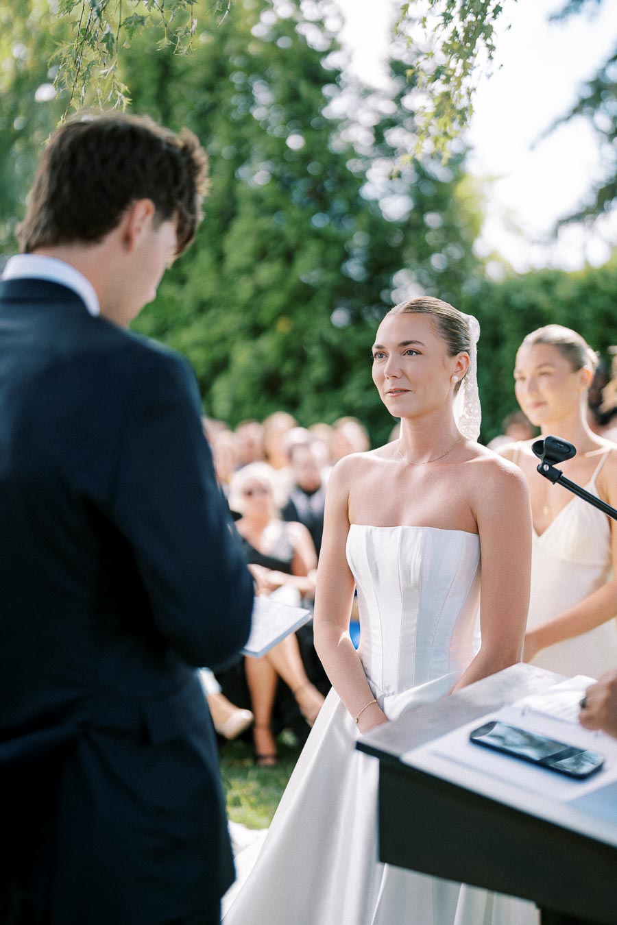 Outdoor wedding ceremony with a bride in a white strapless gown exchanging vows with groom in a suit, surrounded by guests in a lush garden setting.