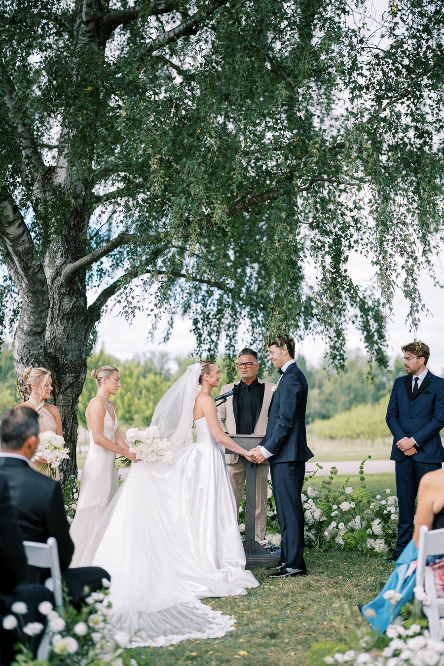 Outdoor wedding ceremony under a large tree, featuring a bride in a white gown and veil holding hands with a groom in a navy suit. Bridesmaids in light dresses stand nearby, with a person officiating the ceremony. Guests are seated, surrounded by lush greenery and floral arrangements.