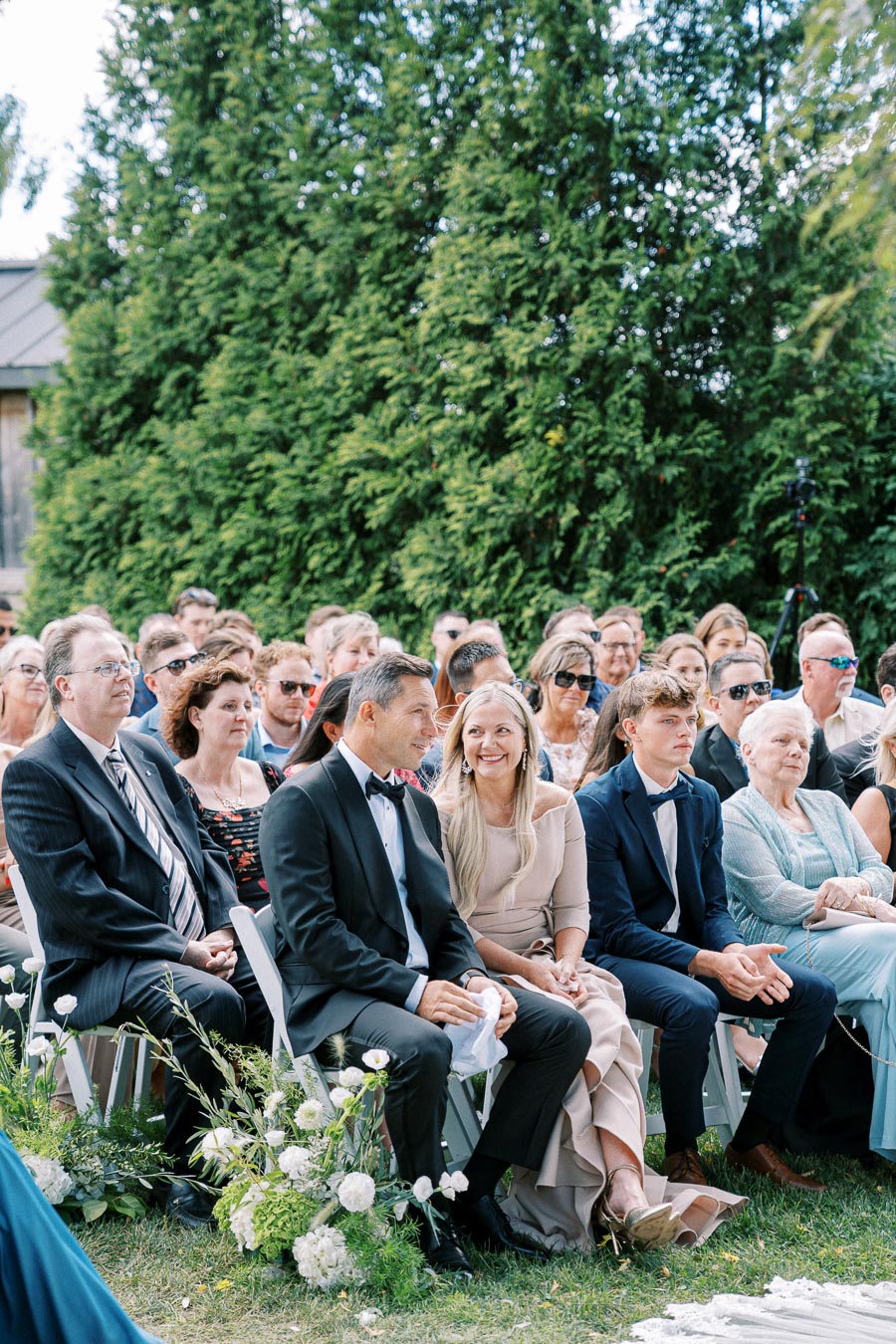 Guests seated at an outdoor wedding ceremony, elegantly dressed with greenery and flowers surrounding them, enjoying the event on a sunny day.