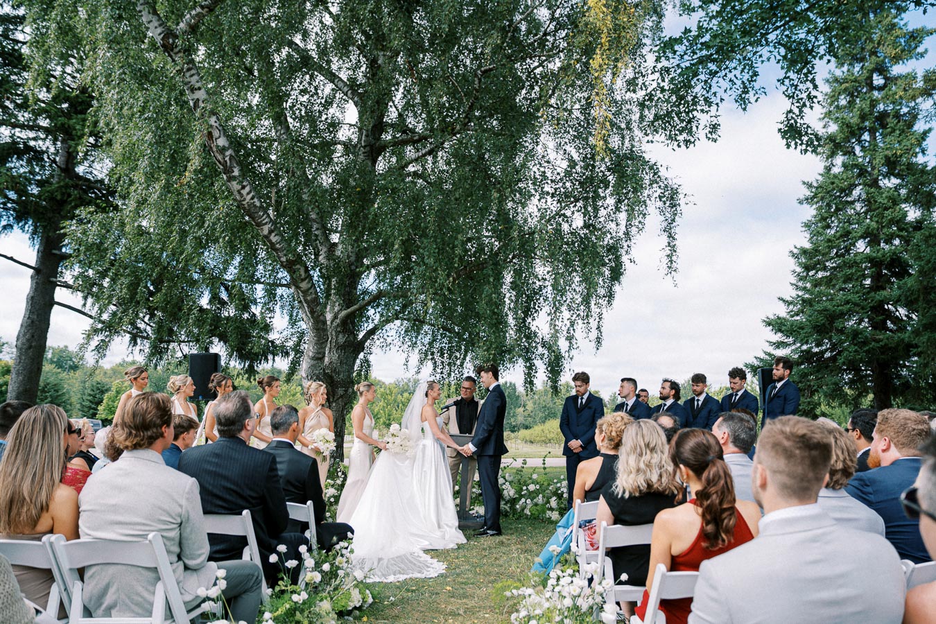 Outdoor wedding ceremony with a bride and groom exchanging vows under a large tree, surrounded by bridesmaids in white dresses and groomsmen in dark suits, with seated guests in the foreground.