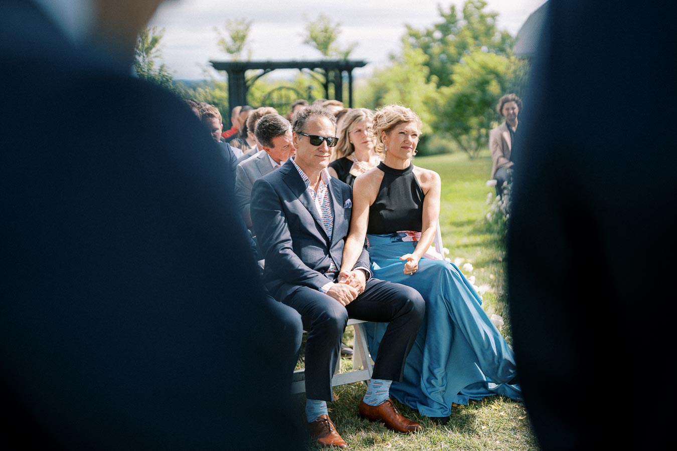 A well-dressed couple sits outdoors at a summer wedding ceremony, surrounded by guests against a backdrop of green trees and a sunny sky.