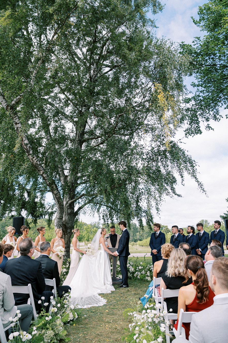 Outdoor wedding ceremony under a large tree, with a bride and groom exchanging vows surrounded by bridesmaids in light dresses and groomsmen in dark suits, attended by seated guests on a sunny day.