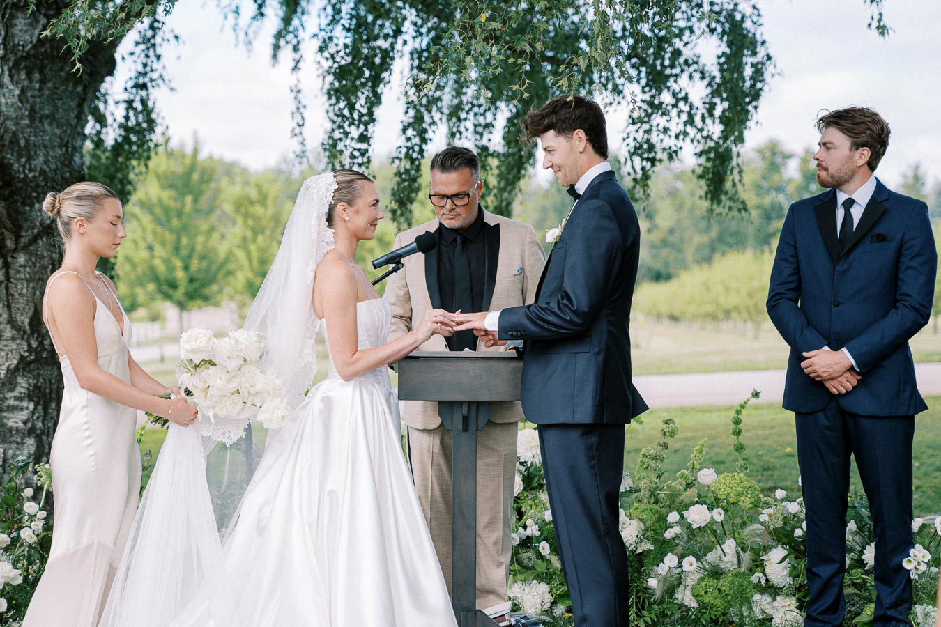 Elegant outdoor wedding ceremony with bride and groom exchanging vows, surrounded by bridesmaids and groomsmen, set against a backdrop of lush greenery.