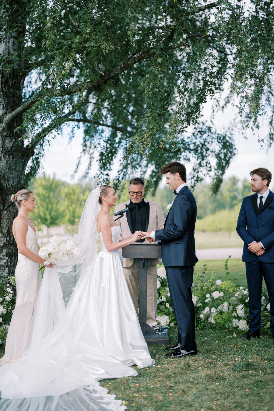 Elegant outdoor wedding ceremony under a large tree with bride in a white gown and veil exchanging vows with groom in a navy suit, accompanied by bridesmaid in a light dress holding a floral bouquet and officiant in the background.